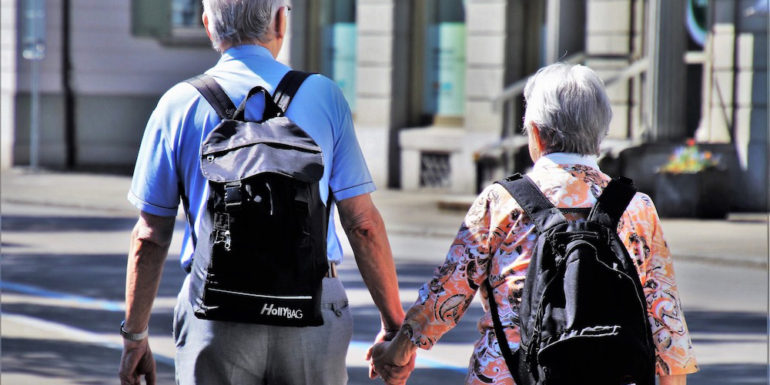 Elderly couple holding hands, each with a backpack.